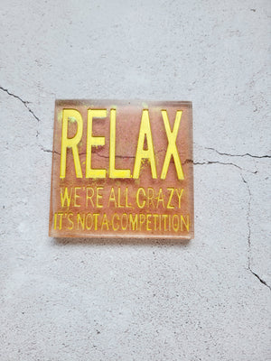 coaster sign made from resin.  It's a clear gold sparkle square with golden letters that says "Relax we're all crazy, it's  not a competition"