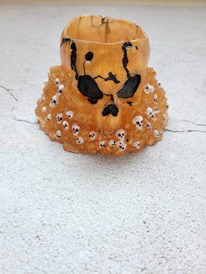 Resin skull dice cup. Big skull holds 2 sets of ttrpg dice comfortably with room for a few extra. The large skull sits on a pile of stones and smaller skulls. The color is a gold with smaller white skulls, black painted eyes, nose, and cracks. Front view