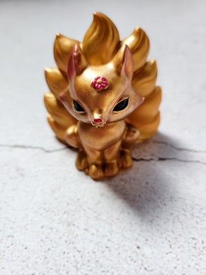 A front view of a kitsune fox figure with nine tails. It's gold in color with a pinkish red flower on its head, black painted eyes and pink painted nose.