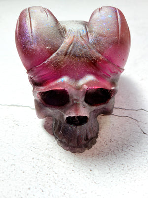 A front view of a skull figure with curved horns on the top of its head. It's darker on the bottom and lighter pinkish red on the top. There are flecks of glitter throughout. It has sunken eyes and nose, textured teeth.