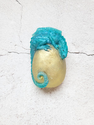 A yellowish gold egg with a teal/turquoise dragon wrapped around on top, its tail curling near the bottom. 