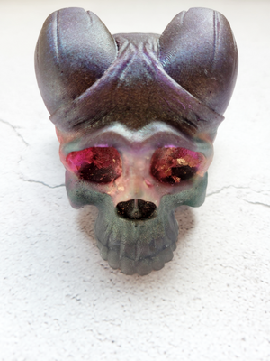 A front view of a skull with curved horns. It's layered with colors of green purple, pinks, and gold flakes.