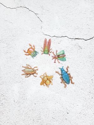A top view of six colorful insect figures. A beetle with green and orange, a cicada with green body and gold wings, a wasp with green wings and pink body, a beetle with blue  body and gold legs, a golden bee, a golden beetle.