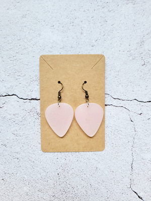 A pair of guitar pick earrings, hanging on a cardboard backer card. The earring hooks are antique toned. The guitar picks are delicate pink with fine gold glitter.