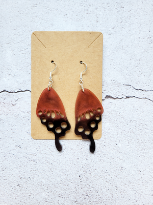 A pair of earrings in the style of butterfly wings on a cardboard backer card. The hook earrings are silver. The wings are dark orange and black.