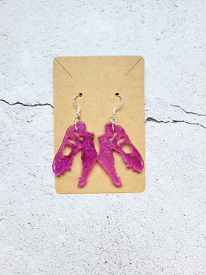 A pair of earrings in the style of t-rex skulls on a cardboard backer card. The hook earrings are silver in color. The skulls are pinkish purple.