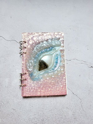 A top view of a closed dragon eye notebook/journal. It has two three-ring clasps in silver. It's filled with A5 paper. The cover is textured. There is a black iris, blue eye scales and pink scales.