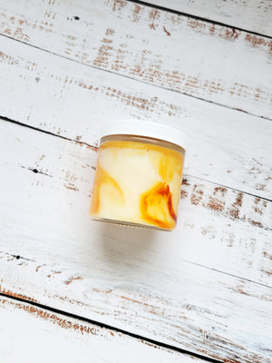 A orange marbled 12oz candle laying on its side to show the swirls of the color. It has a white lid and a glass jar. Candle name is Supernatural: "Pudding!". Scent profile is french vanilla and butterscotch.