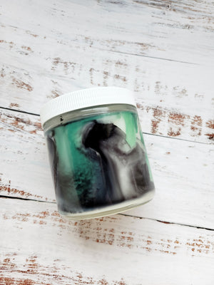 A side view of a 12oz marbled candle to show the black and green swirls in the wax. It's in a glass jar with a white lid. This candle is called Wild Magic Surge: Shrine. It's scent profile is smoke, incense, vanilla. It has a mystery Dungeons and Dragons die inside.