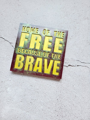 Square coaster sign that reads "Home of the free because of the brave". it's a reddish color with gold text