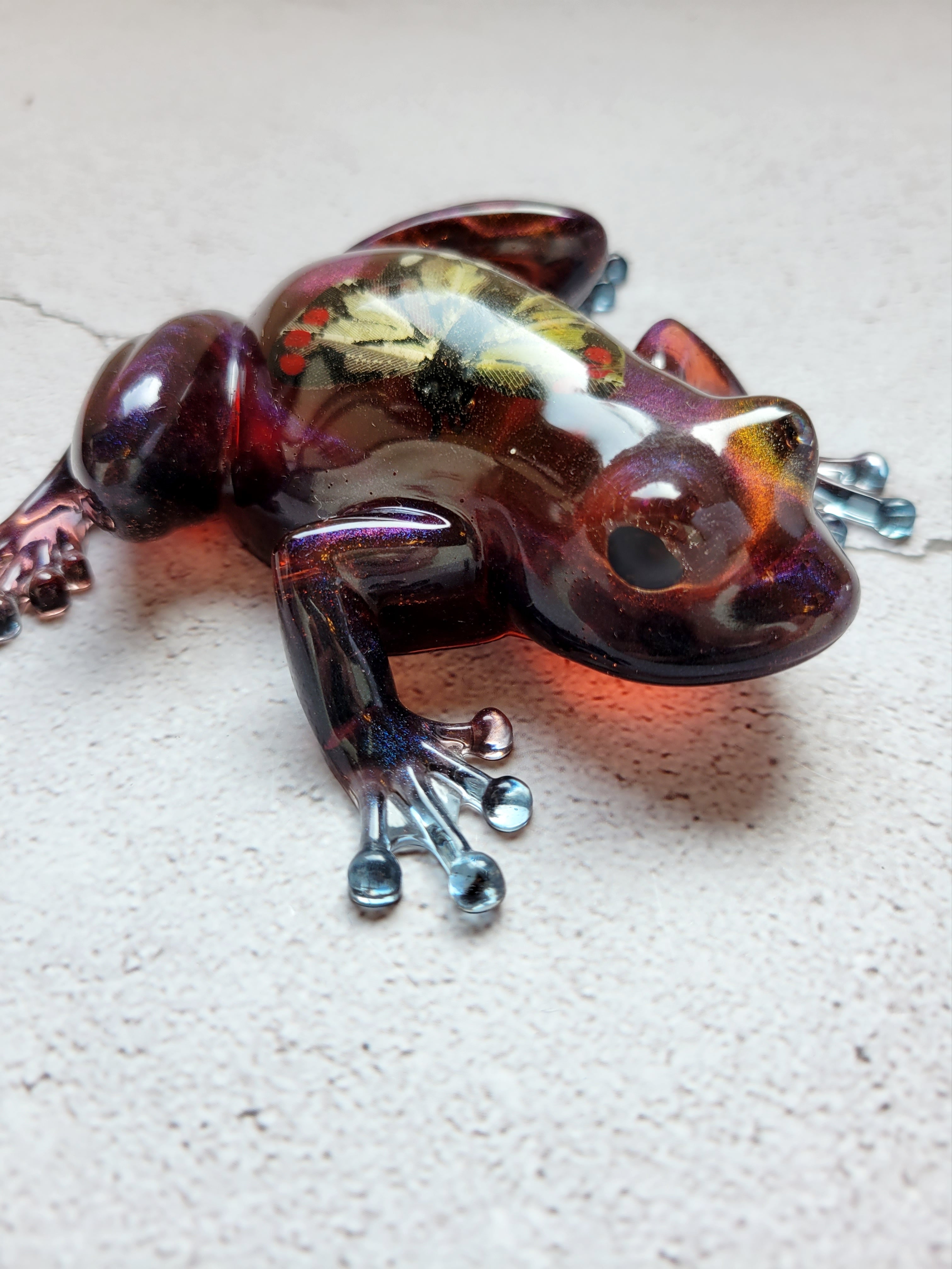 frog resin with a paper yellow butterfly inside. The frog is color shifting with blues and reds and purples. Front face view