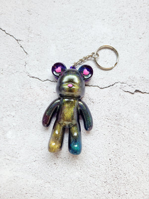 Top view of a snow goggle wearing bear keychain with pink nose and heart ears. The color is color shifting golds, blues, purples, with a silver keychain hoop