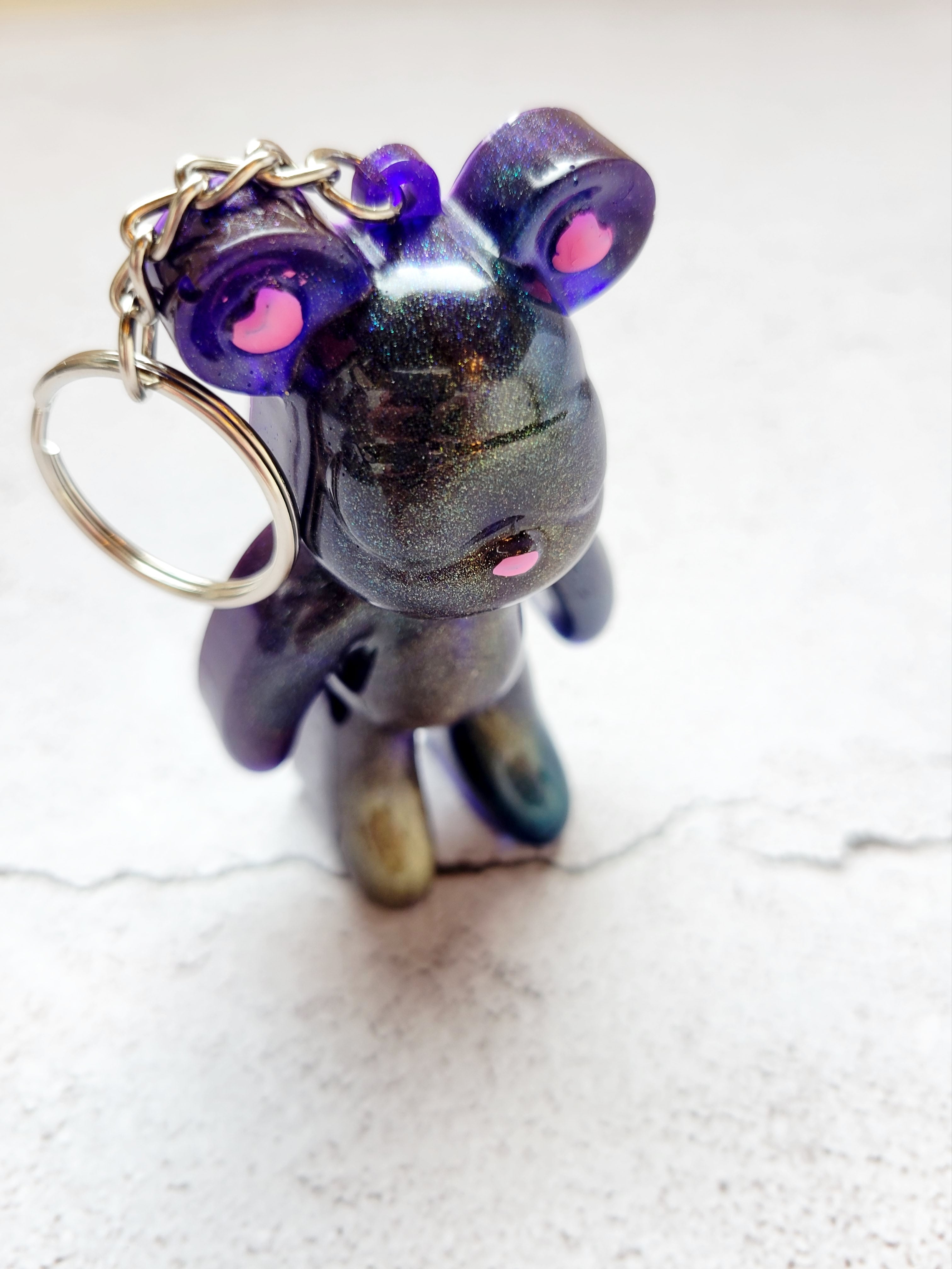 Standing front view of a snow goggle wearing bear keychain with pink nose and heart ears. The color is color shifting golds, blues, purples, with a silver keychain hoop