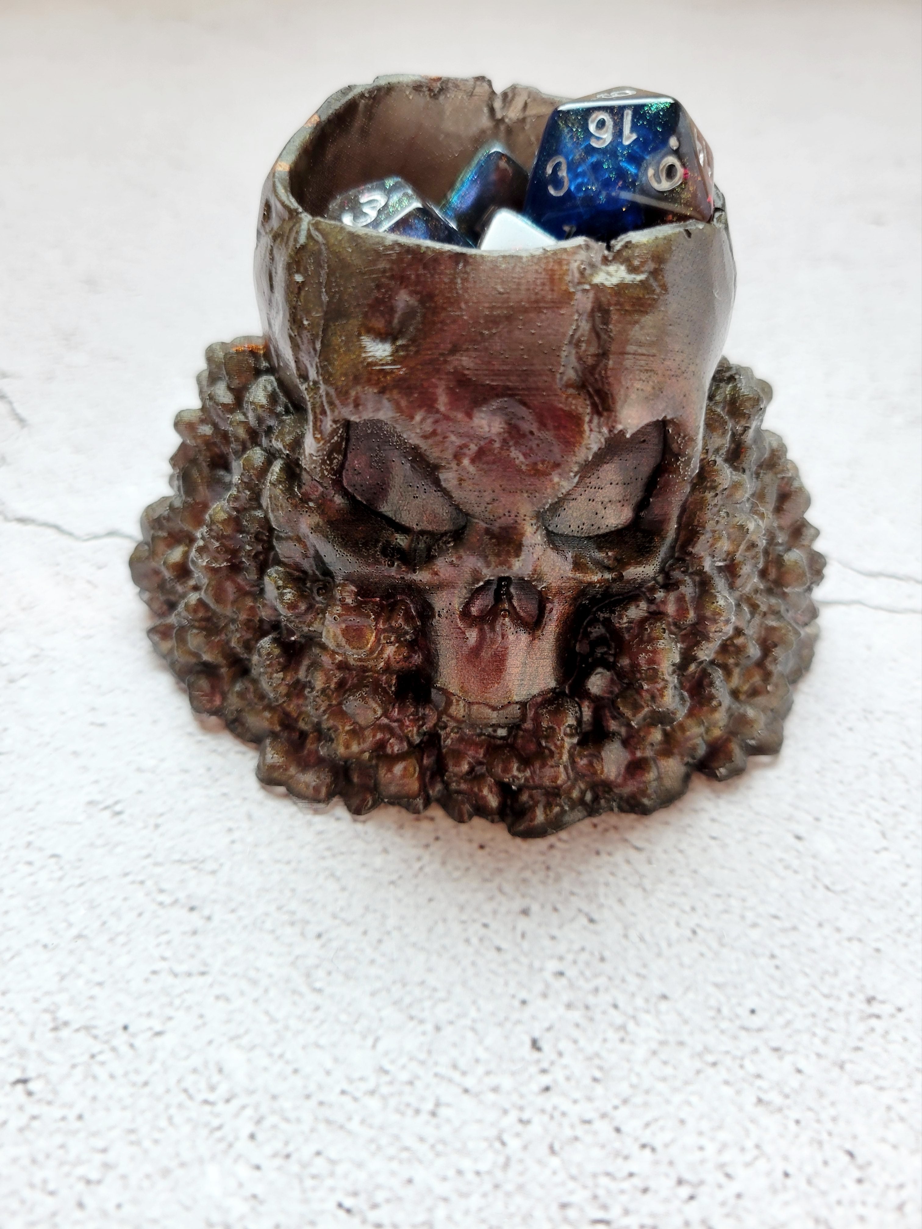 Resin skull dice cup. Big skull holds 2 sets of ttrpg dice comfortably with room for a few extra. The large skull sits on a pile of stones and smaller skulls. The color is a bronze reddish brown. Front view with dice inside