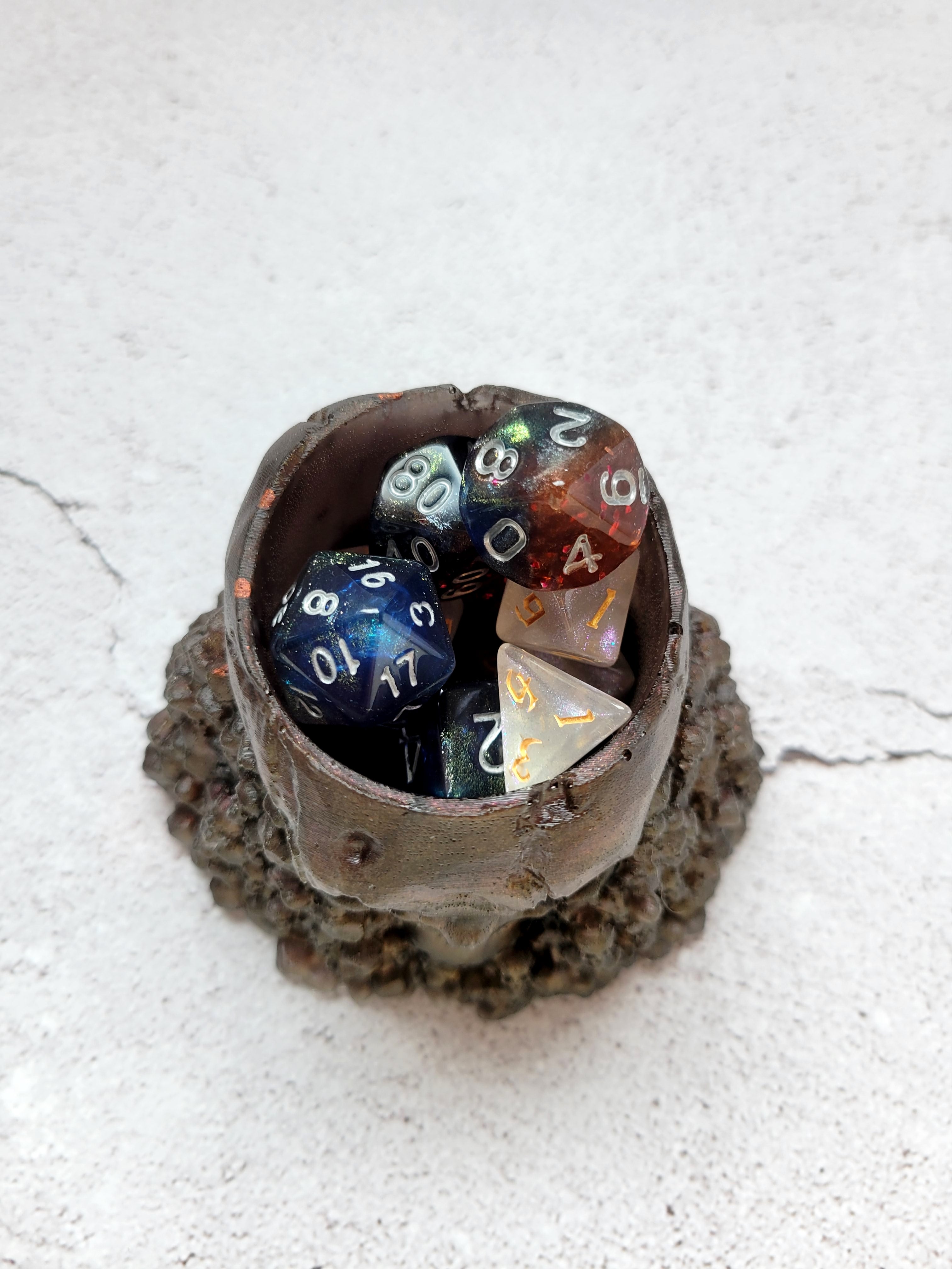 Resin skull dice cup. Big skull holds 2 sets of ttrpg dice comfortably with room for a few extra. The large skull sits on a pile of stones and smaller skulls. The color is a bronze reddish brown. top view with dice inside