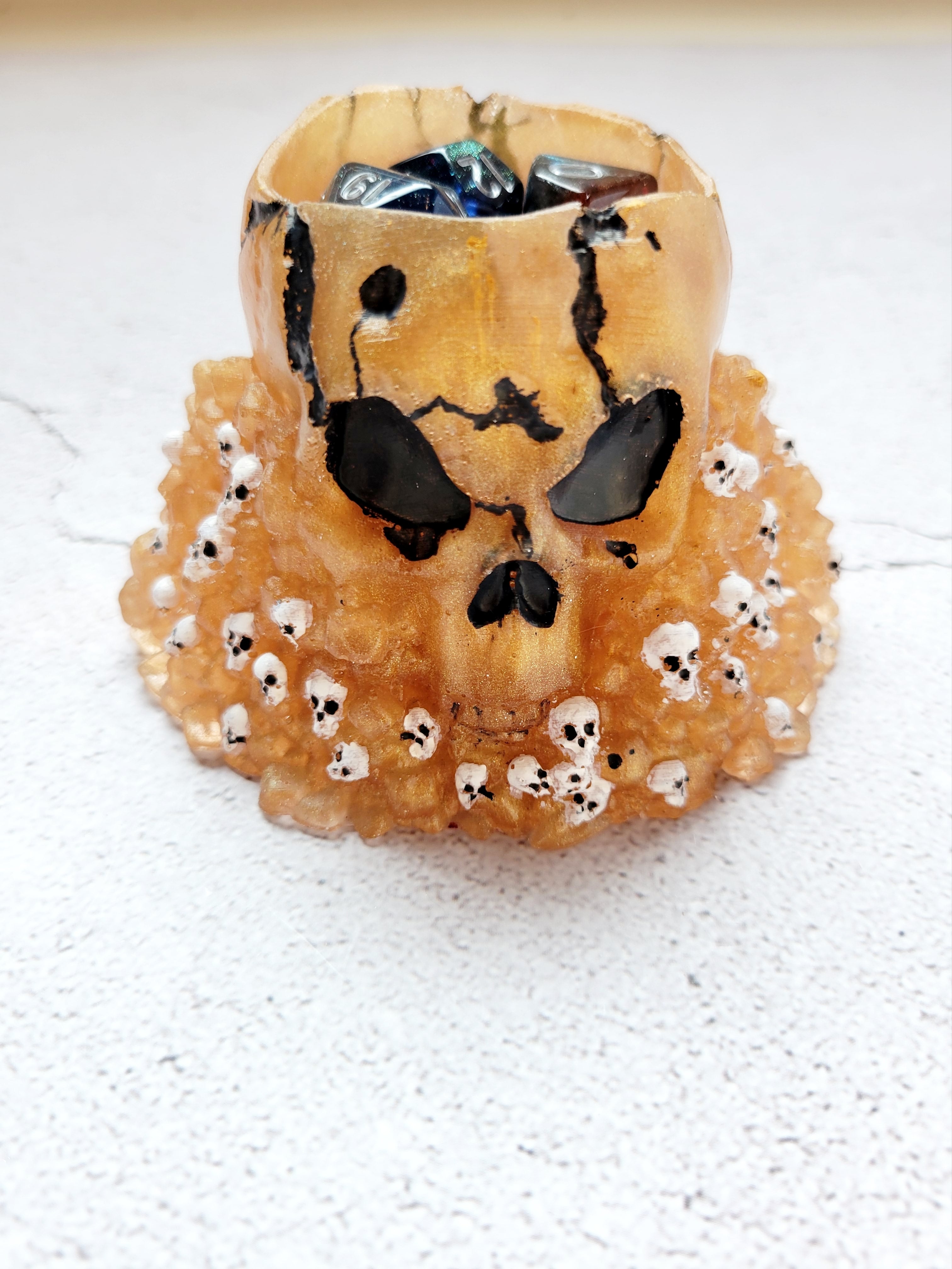 Resin skull dice cup. Big skull holds 2 sets of ttrpg dice comfortably with room for a few extra. The large skull sits on a pile of stones and smaller skulls. The color is a gold with smaller white skulls, black painted eyes, nose, and cracks. Front view with dice inside