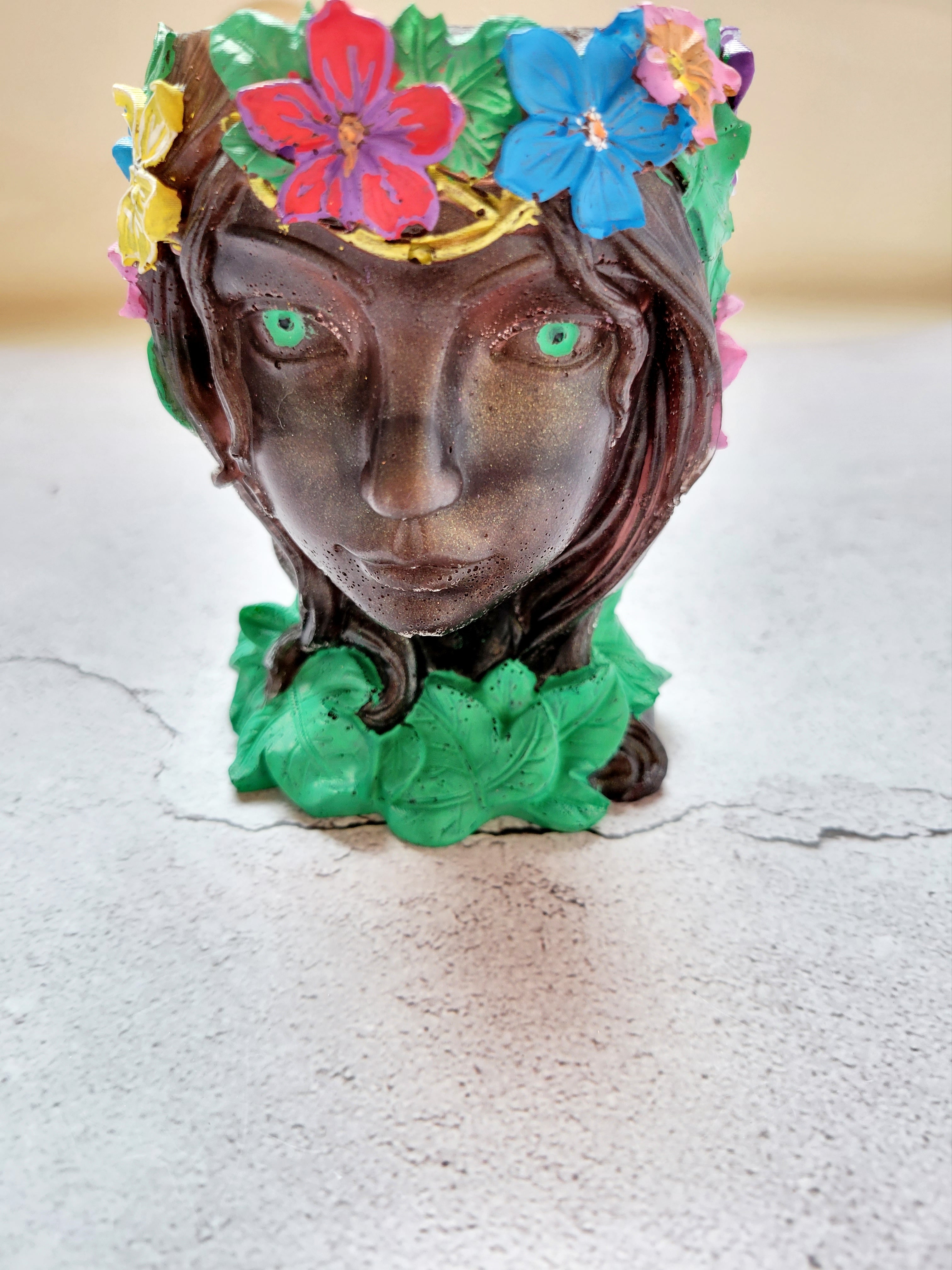 brownish bronze colored resin woman's face with green eyes, green ivy around her neck, a golden stem crown filled with colorful flowers. Front view