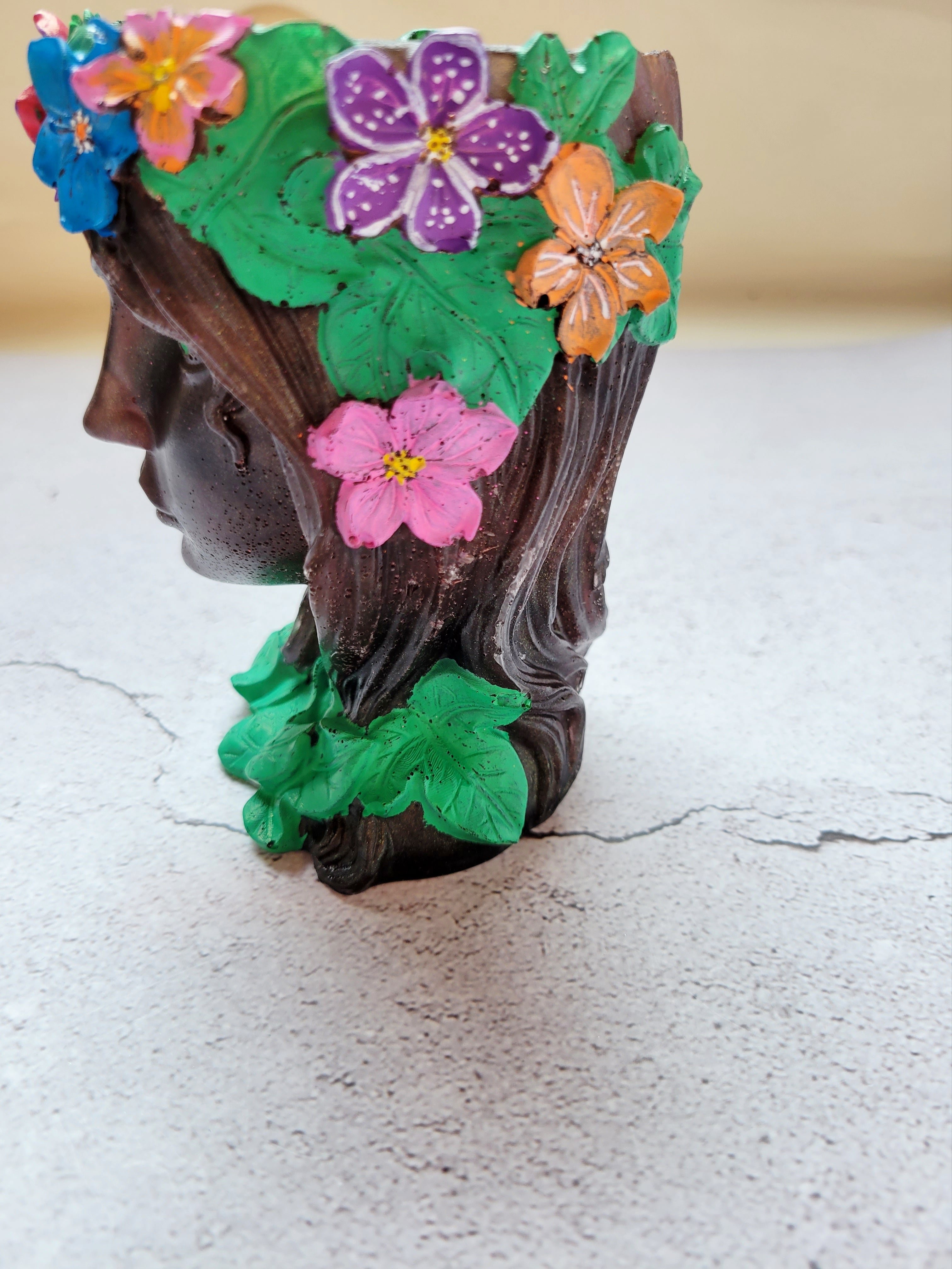 brownish bronze colored resin woman's face with green eyes, green ivy around her neck, a golden stem crown filled with colorful flowers. Side view