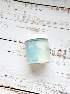 A 12oz marbled candle laying on its side to show the pale blue swirls in the wax. It has a white lid and glass jar. This candle is called Wild Magic Surge: Bathhouse. It's scent profile is teak, bamboo, and flower petals. It has a mystery Dungeons and Dragons die inside.