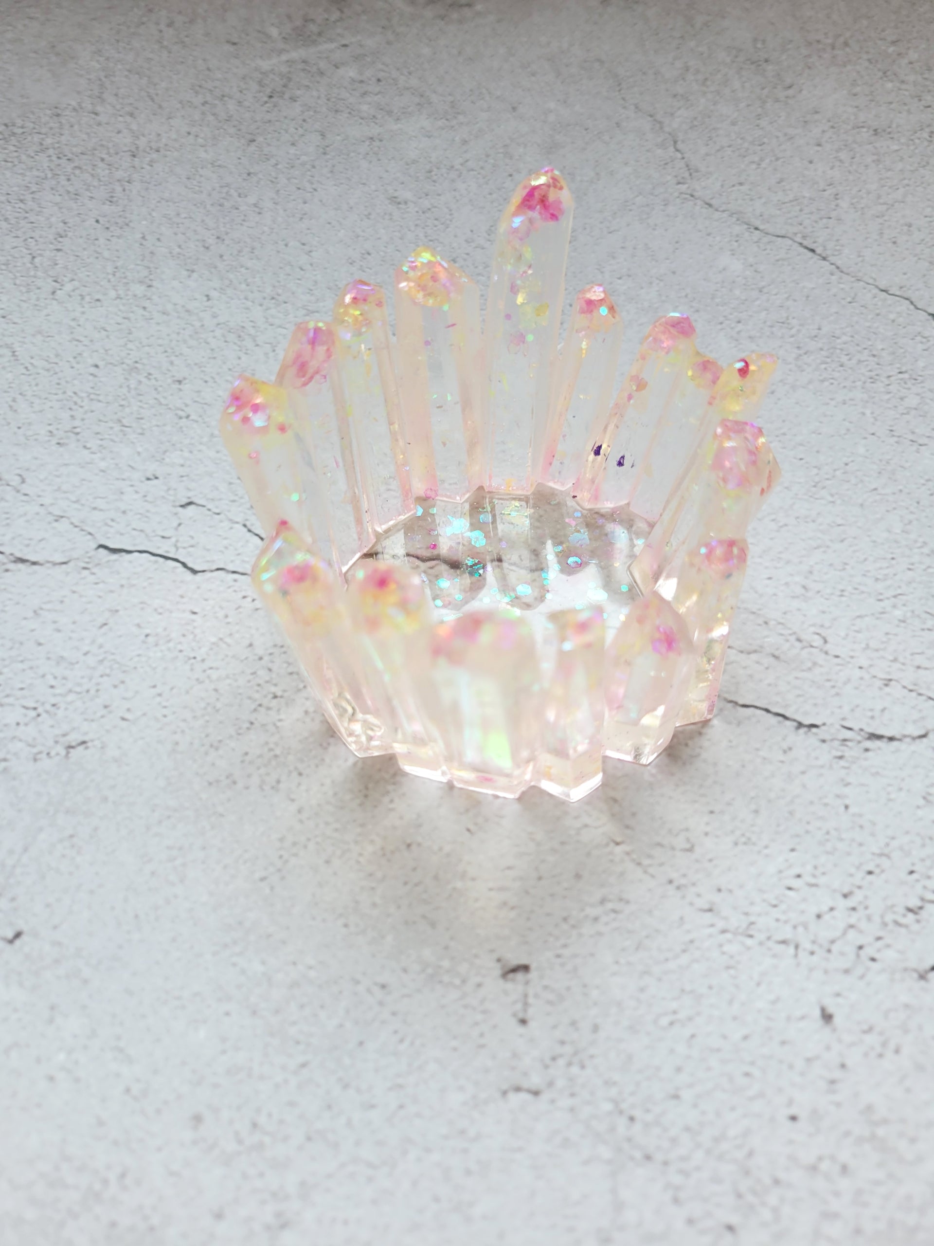 A side birds eye view of a tealight candle holder in the style of a cluster of crystals forming a circle. It's translucent clear with pink and yellow glitter. The crystals are various heights.