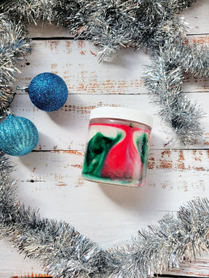 A side view of a 12oz marbled candle to show the red and green swirls in the wax. It's in a glass jar with a white lid. It's surrounded by holiday decorations. This candle is called Holiday Special: Trimming the Tree. It's scent profile is pine, orange, cranberry.