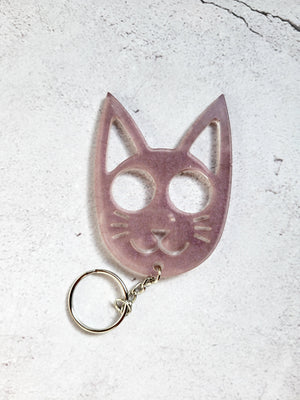 A defense keychain in the style of a cat face. It has a silver chain and ring hoop. It's purple in color. 