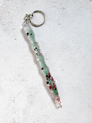 A defense keychain with a silver chain and ring hoop. It's a pale green color with various glitter sizes and colors throughout.