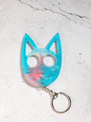 A defense keychain in the style of a cat face. It has a silver chain and ring hoop. It's a light blue with pink patch.