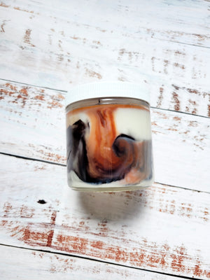 A 12oz marbled candle laying on its side to show the brown and black swirls in the wax. It has a white lid and glass jar. This candle is called Timeless: Christopher's Bunker. It's scent profile is leather, cedar, incense.