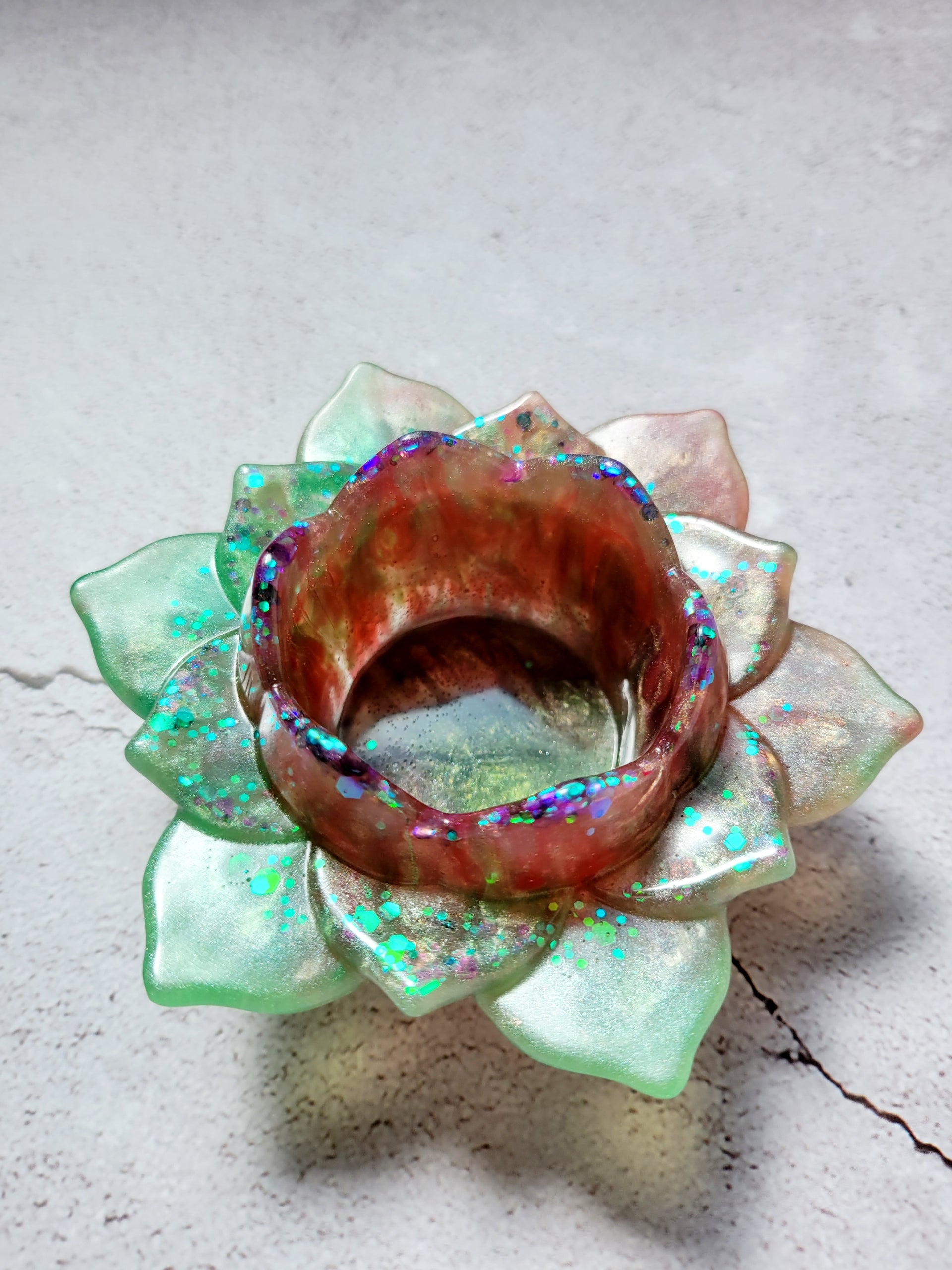 A side birds eye view of a tealight candle holder in the style of a lotus flower. It's got green and brown coloring with flecks of colorful glitter along the petals. 