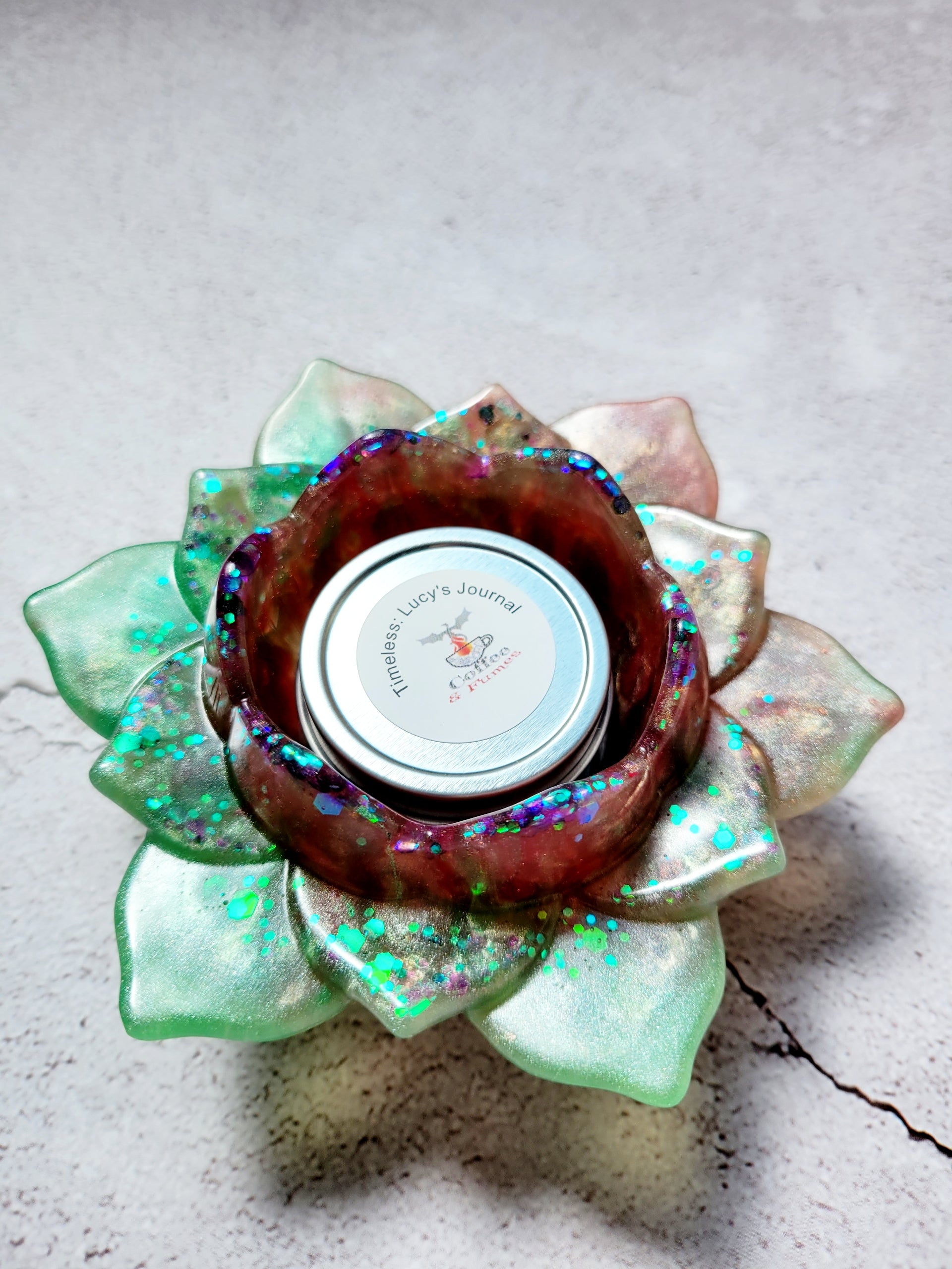 A side birds eye view of a tealight candle holder in the style of a lotus flower. It's got green and brown coloring with flecks of colorful glitter along the petals.  There is a tealight candle inside to show size comparison.