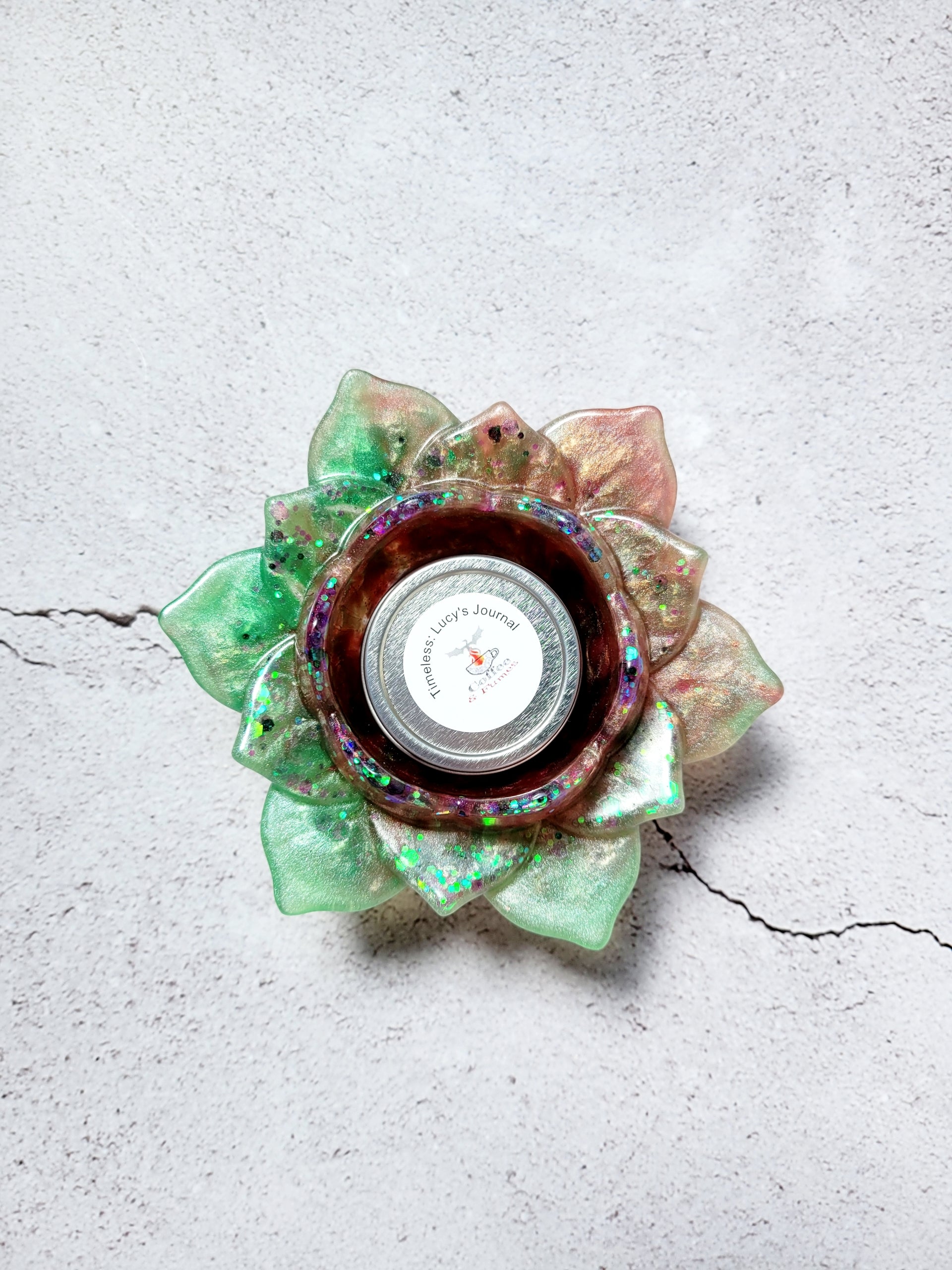 A birds eye view of a tealight candle holder in the style of a lotus flower. It's got green and brown coloring with flecks of colorful glitter along the petals.  There is a tealight candle inside to show size comparison.