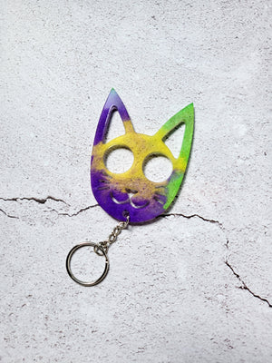 A cat face defense keychain with pointy ears, cutout eyes for fingers, cutout whiskers and mouth. It's got a silver chain and hoop below the mouth. Its purple, yellow, and lime green in color. 