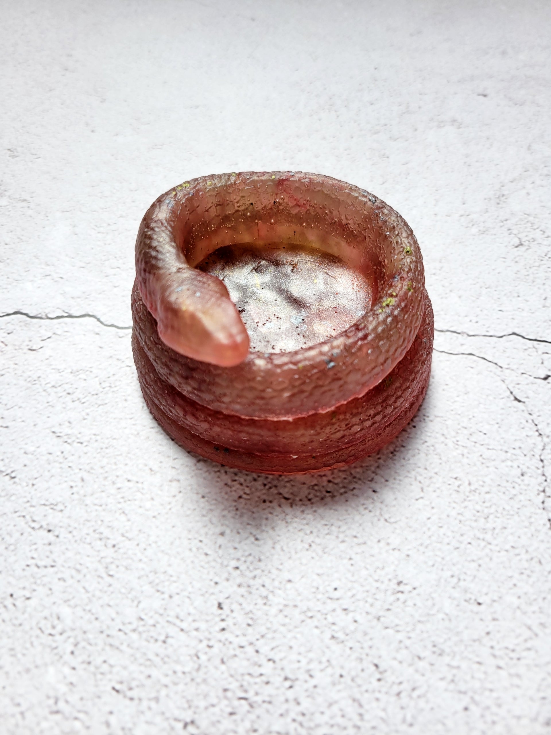 A side view of a tealight candle holder in the style of a coiled snake. It's brownish red in color with flecks of yellows and grays. 