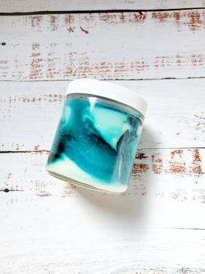 A 12oz marbled candle laying on its side to show the teal blue swirls in the wax. It has a white lid and glass jar. This candle is called Critical Role: Dorian's Bacon Lips. It's scent profile is bacon, amber, vetiver. It has a mystery Dungeons and Dragons die inside.