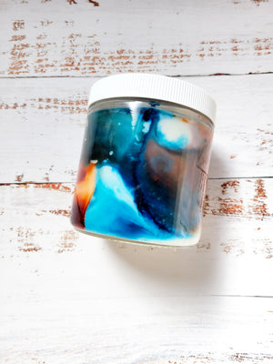 A 12oz marbled candle laying on its side to show the blue and brown swirls in the wax. It has a white lid and glass jar. This candle is called Critical Role: Beau's Pocket Bacon. It's scent profile is bacon, maple, black pepper. It has a mystery Dungeons and Dragons die inside.