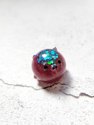 A small round pig figure with black painted eyes and nose. it's a deep purple red color with blueish green glitter on the top of its head.