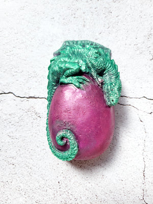A dragon with egg figure. The egg is a purple red color. The dragon is perched on top, its tail wrapping around the bottom. The dragon is green. 