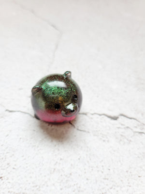 A small round bear figure with black painted eyes and nose. He's a color shifting green and red. 