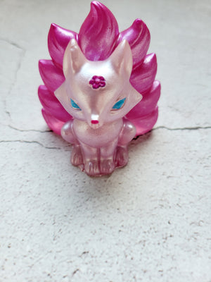 A front view of a kitsune fox figure with nine tails. It has a pale pink body with dark pink tails. It's eyes are blue and nose is pink. The flower on its head is the color of the tails.