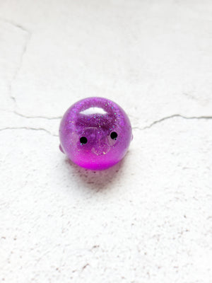 A small round penguin figure with black painted eyes. It's a bright purple color with fine glitter throughout.
