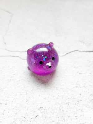 A small round bear figure with black painted eyes and nose. It's purple with glitter.