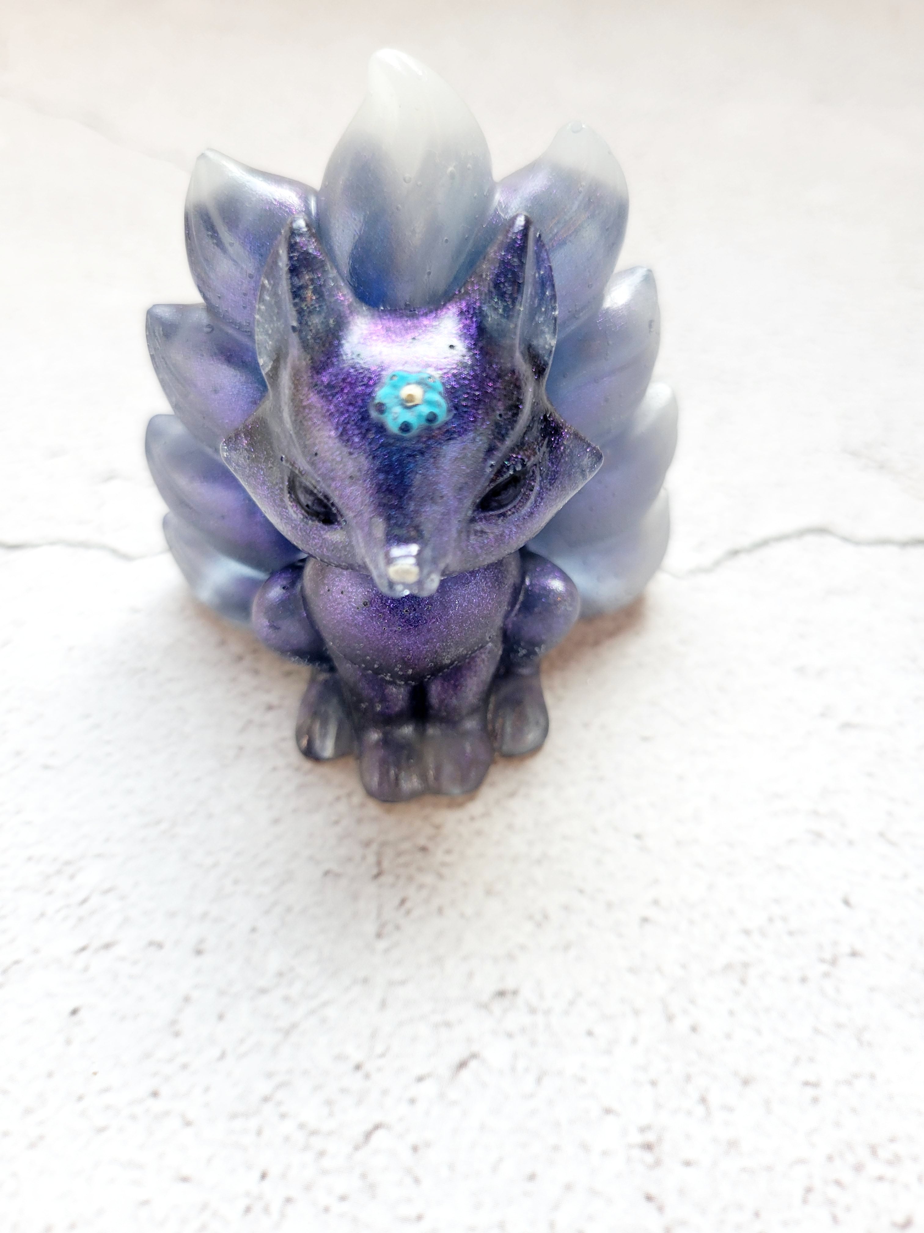 A front view of a kitsune fox figure with nine tails. It's body is deep blue with a blue and white swirled tail. It has black eyes, white nose, and light blue flower on its head.