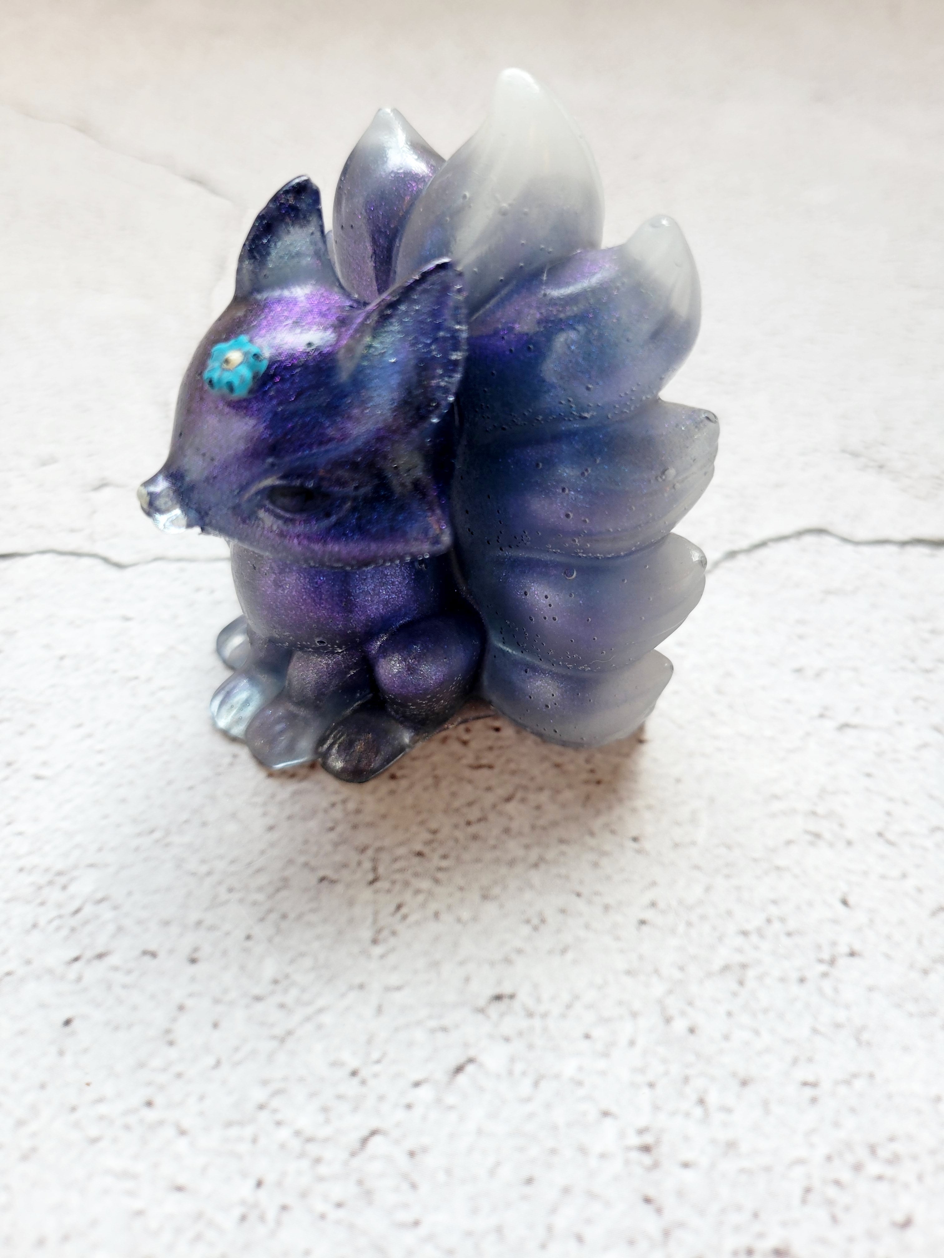 A side view of a kitsune fox figure with nine tails. It's body is deep blue with a blue and white swirled tail. It has black eyes, white nose, and light blue flower on its head.