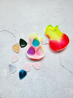 A top view of an acoustic guitar body - a case for guitar picks. It holds nine randomly colored guitar picks. The case is red, yellow, pink, white. The picks are varied pinks, blues, blacks, golds. It's open to show everything.