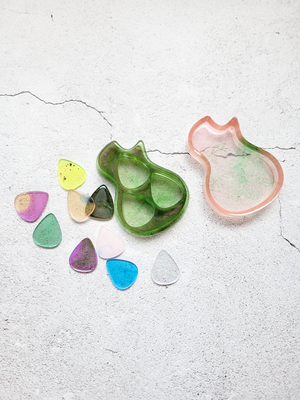 A birds eye view of a guitar pick case in the style of an acoustic guitar body. It's open with nine various guitar picks laying around it. The lid is a translucent pink with hints of green. The bottom is green with hints of pink. There are three pick sized holes in the base. The picks are a variety of colors: black, yellow, pink and gold, green, blue, clear.