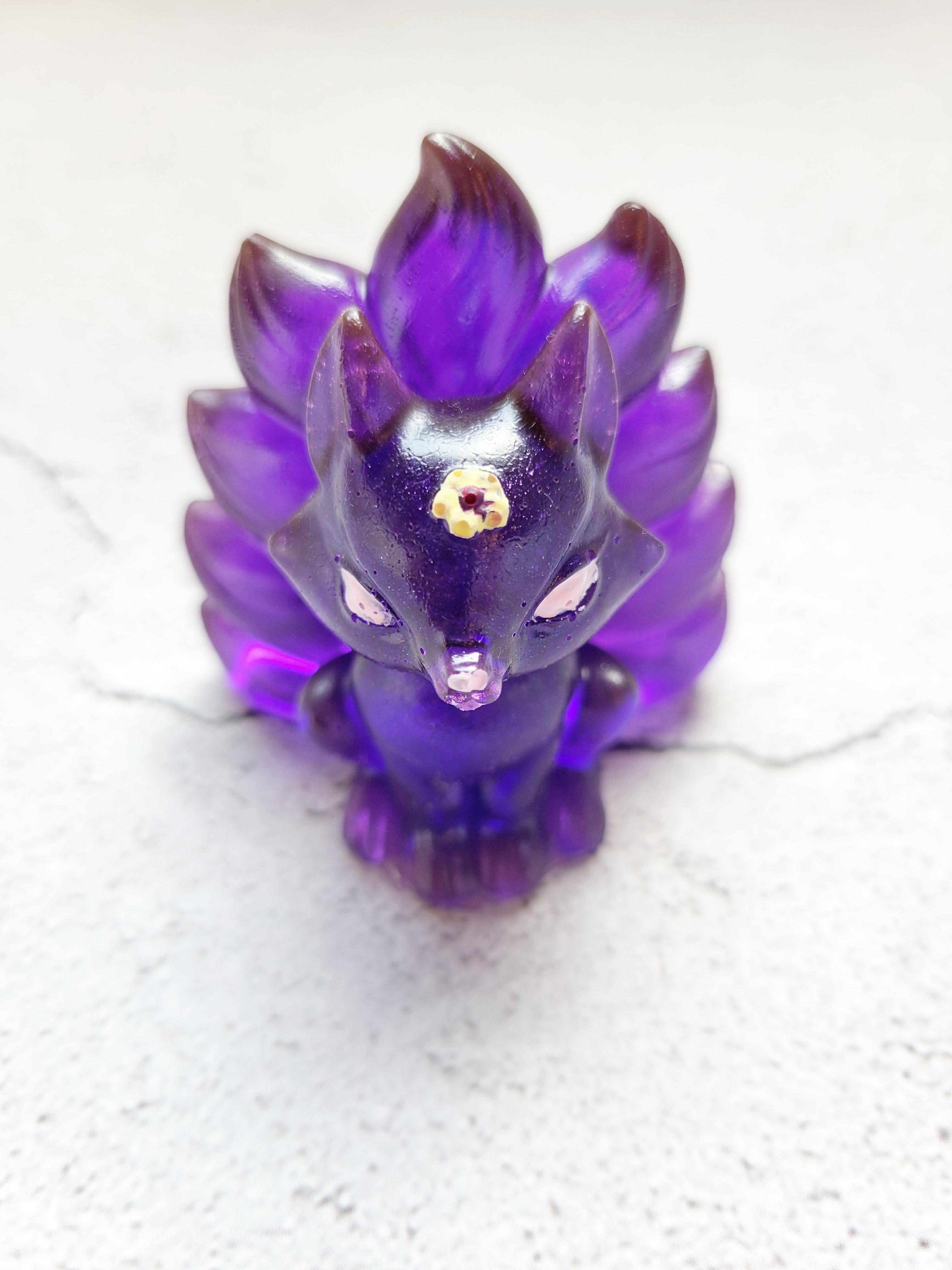 A front view of a sitting kitsune fox figure with nine tails. It's a deep purple color with a yellow flower on its head, white eyes and nose.