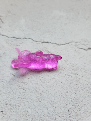 A side view of a small sleeping fox figure stretched out on its back. It's a bright transparent pink with fine glitter throughout with black painted eyes and nose.