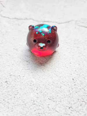 A small round bear figure with black painted eyes and nose. It's red with some blue and green glitter sitting on top of its head.