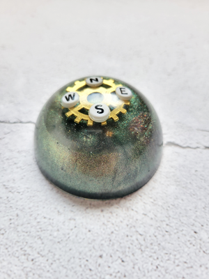A side view of a tabletop compass. There's a gold gear with the direction letters, N, E, S, W, on it. It's various shades of green, gold, red.